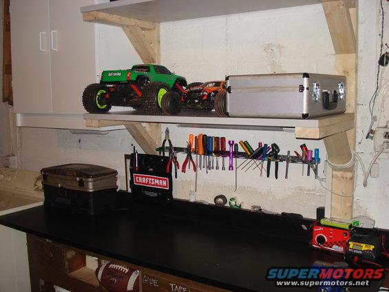 Green and orange remote control monster trucks on a shelf above a workbench with tools