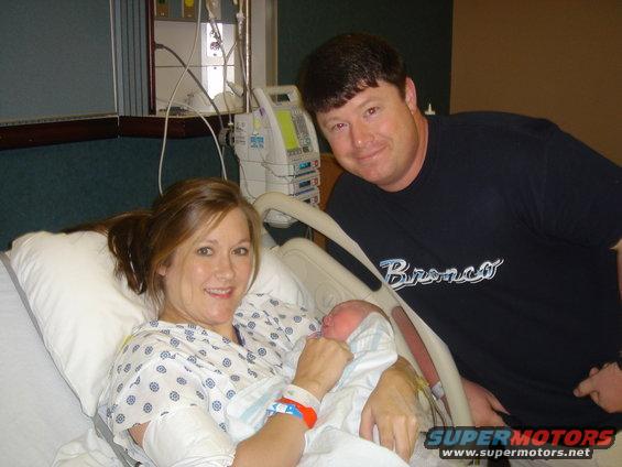 Mom and dad with their newborn baby in a hospital bed. Dad wears a shirt that says 'Bronco'