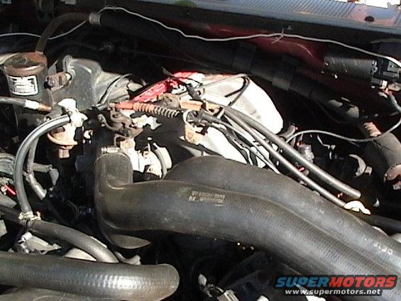 Engine bay with hoses, battery, and wiring visible. Some text is visible on a black hose