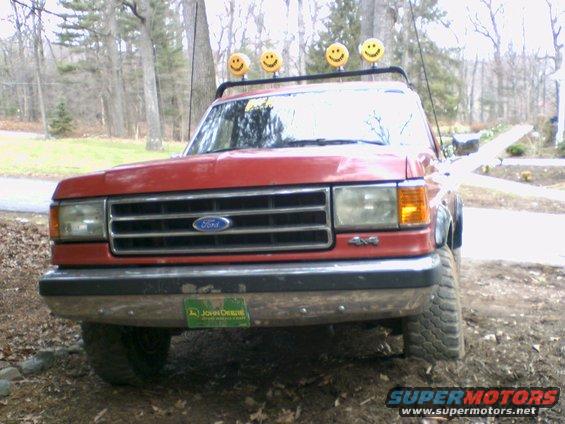 Front view of a red Ford pickup with a John Deere plate and smiley face lights on the roof rack