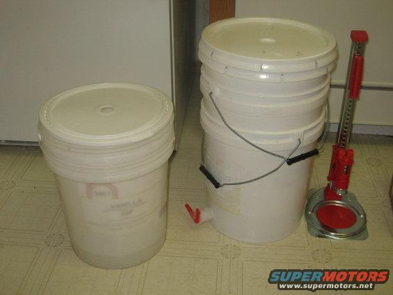 Two white buckets, one with a spigot, and a red bottle jack on a tiled floor