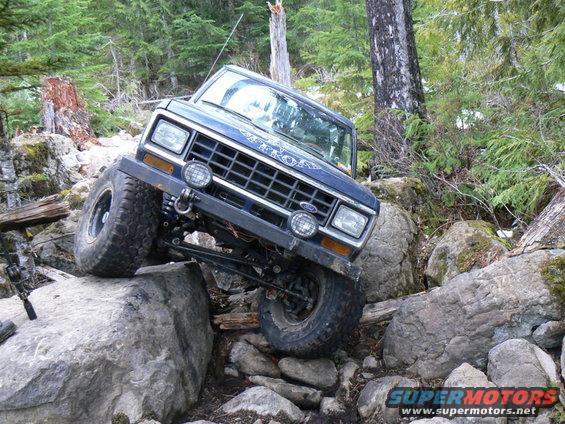 Blue 1986 Ford Bronco II climbing over large rocks on a wooded trail