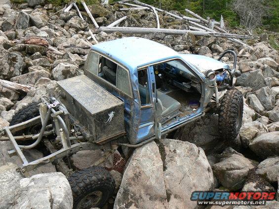 Blue off-road vehicle with its doors removed is wedged between large rocks on a wooded trail
