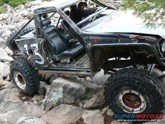 Heavily modified off-road vehicle with exposed interior and roll cage, navigating rocky terrain