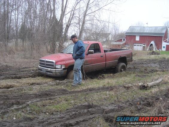 Red Dodge Ram pickup truck stuck in mud with a man standing beside it. A red barn is in the background