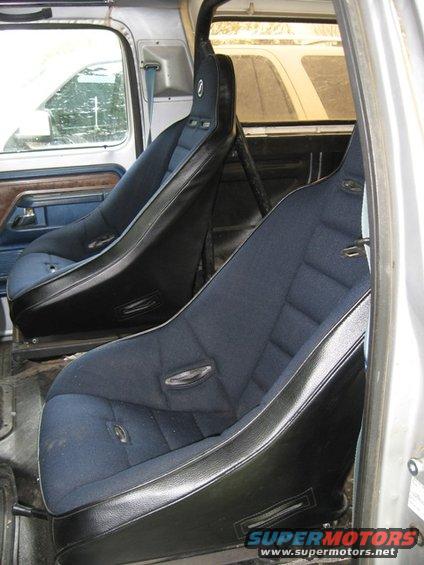 Interior view of a vehicle showing two dark blue bucket seats with black trim and a roll cage. The door panel has wood grain accents