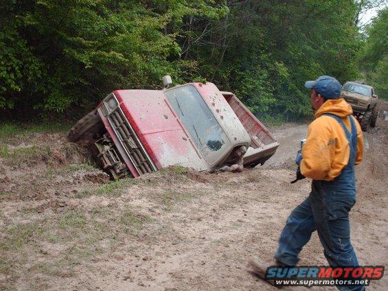 Red pickup truck tipped on its side in mud, with a man in an orange jacket and blue overalls walking away. Another truck is visible in the background