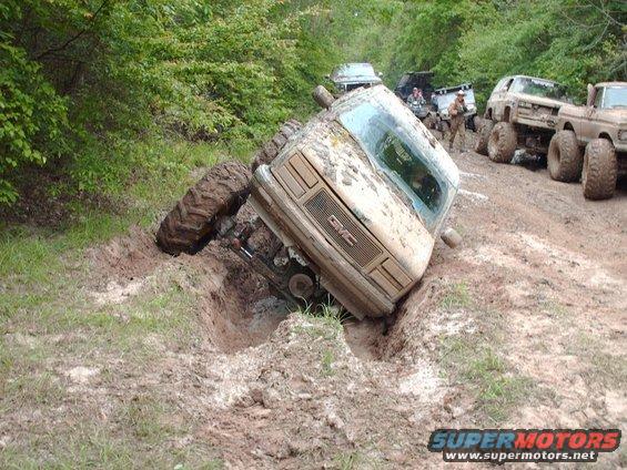Mud-covered GMC truck tilted in a ditch, front grille visible. Other trucks in background