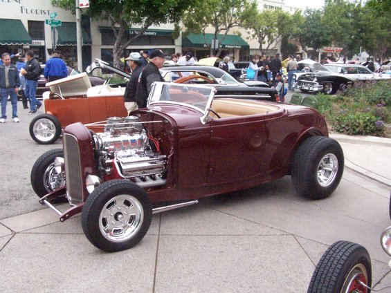 Maroon hot rod roadster with exposed chrome engine and tan interior, seen from a 3q front view at a car show