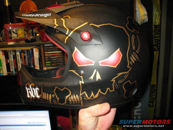 Black motorcycle helmet with red glowing eyes and gold skull graphics, held by a hand