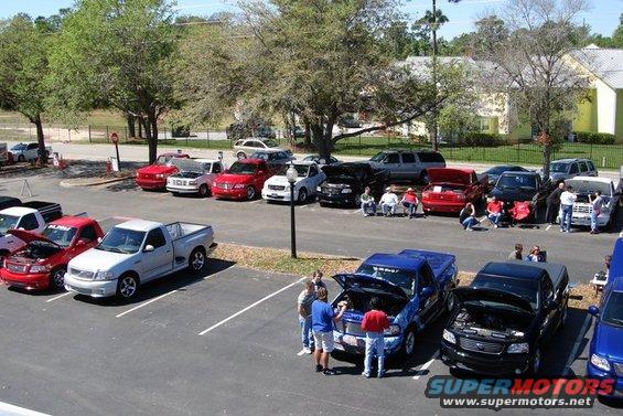 Parking lot gathering with many pickup trucks and cars, some with hoods open