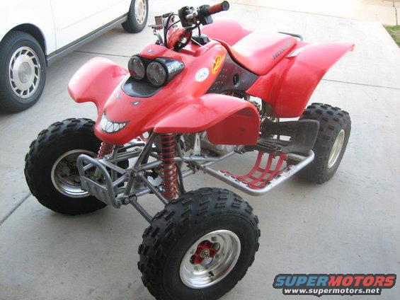 Red ATV with dual headlights and a large, cartoonish grin sticker on the front fairing. A white car is partially visible to the left
