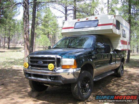 Green Ford F-250 pickup truck with a camper shell in a wooded area. It has yellow smiley face covers on the headlights
