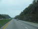 1995 Ford C-350 (Centurion Conversions) - Cars travel on a multi-lane highway bordered by trees and rocky terrain under an overcast sky.