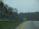 1995 Ford C-350 (Centurion Conversions) - A view from a vehicle traveling on a highway, showing a grassy shoulder and a wooded hillside.
