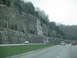 1995 Ford C-350 (Centurion Conversions) - A highway passes by a large rock cliff with trees on top, under an overcast sky.