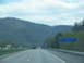 1995 Ford C-350 (Centurion Conversions) - View from a car on a highway approaching a welcome center, with forested mountains under a cloudy sky.