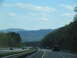 1995 Ford C-350 (Centurion Conversions) - A dark truck travels down a highway surrounded by rolling hills and mountains under a partly cloudy sky.