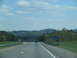 1995 Ford C-350 (Centurion Conversions) - A car drives down a highway with rolling hills and trees in the background under a cloudy sky.