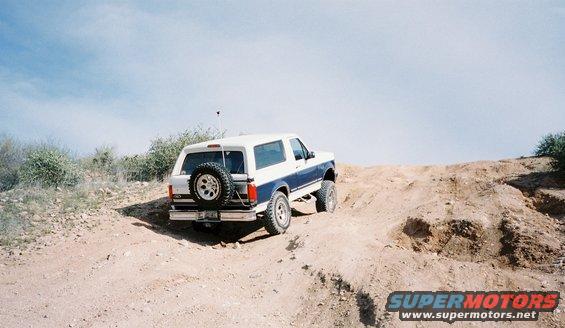 White and blue Ford Bronco climbing a dirt hill with a spare tire on the back