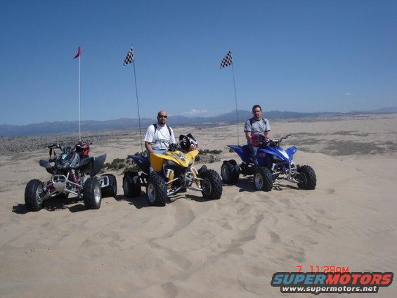 Three ATVs parked on sand dunes with riders, flags flying