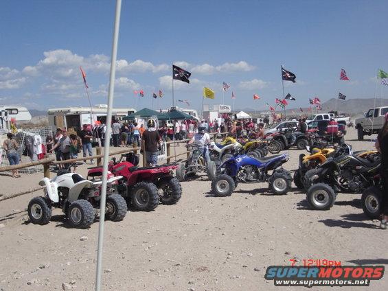 Several ATVs parked on dirt, with people and trailers in the background under a blue sky