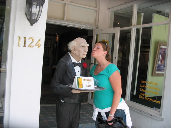 Woman kisses mannequin waiter holding a sign that says "The Gallery Is Open"