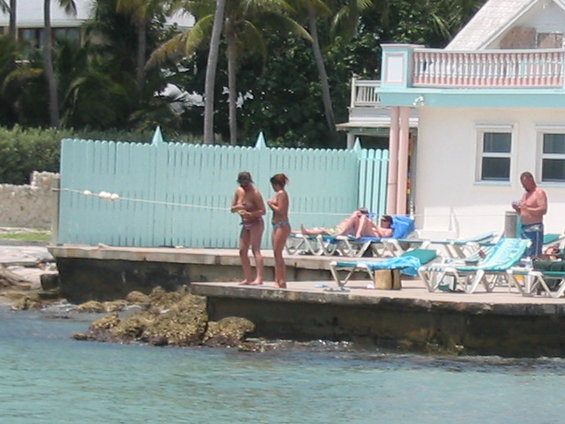 People relaxing on lounge chairs and standing on a boardwalk next to the water, with palm trees and a building in the background