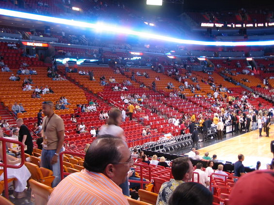 Spectators fill the red and orange seats of a basketball arena, with a court visible in the background