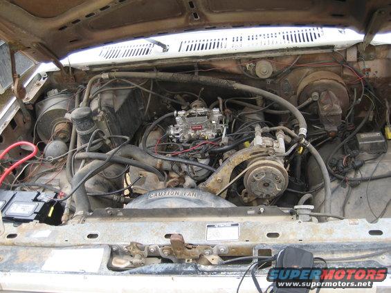 Engine bay with V8 engine, carburetor, and air conditioning compressor. Rusty hood and fan shroud