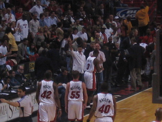 Basketball players in white jerseys with red trim walk on court, crowd in background