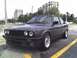 1989 BMW 3 Series - A dark gray 1989 BMW 3 Series coupe is parked in a lot with buildings in the background.
