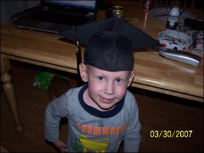 Child in graduation cap and "teamwork" shirt smiles at camera, with a laptop and table in background
