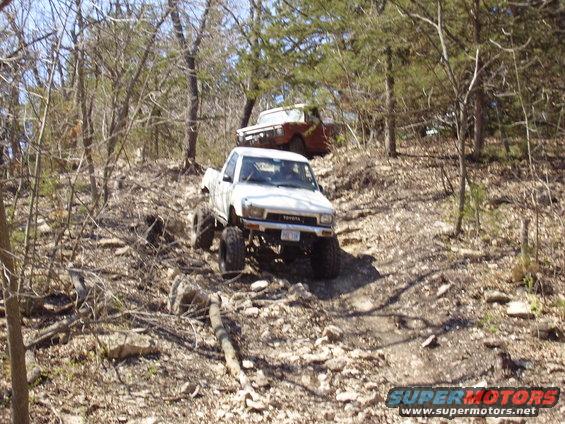 White Toyota pickup truck with large tires climbing a rocky hill, another older truck visible in the background