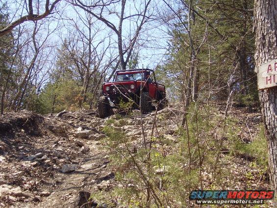 Red off-road vehicle with roll cage climbing a steep, rocky trail in a wooded area