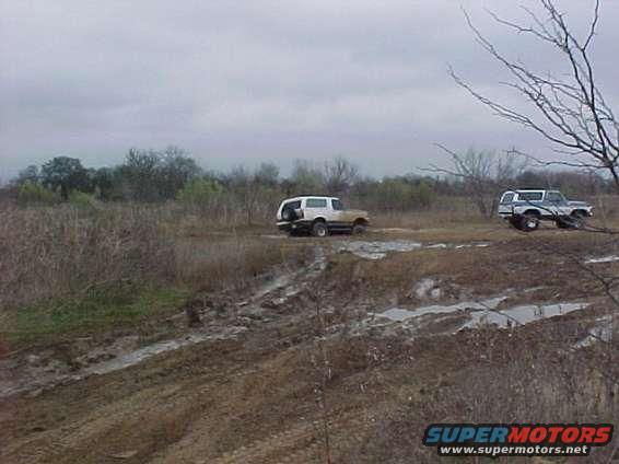 two SUVs driving through a muddy, grassy field under a cloudy sky