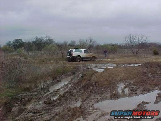 White SUV stuck in mud, rear spare tire visible, cloudy day