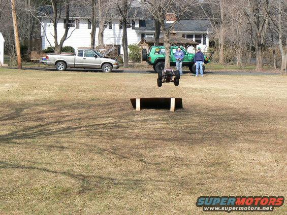 Remote control buggy mid-air over a ramp, with two people and two full-size vehicles in background