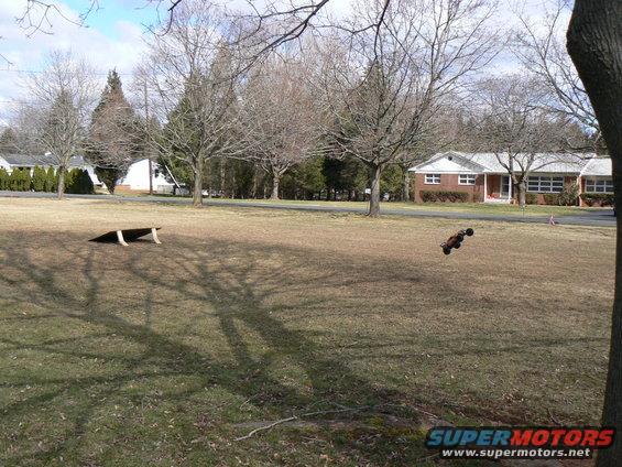 RC car airborne over grassy field with ramp nearby and houses in background