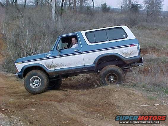 Blue and white 1978 Ford Bronco driving on a dirt trail with trees in the background