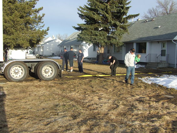 Trailer wheels and several people standing in a muddy yard with houses in the background