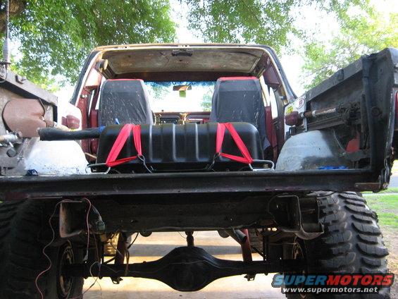 Rear view of a red 1978 Ford Bronco with its tailgate down, showing a black auxiliary fuel tank secured with red straps inside
