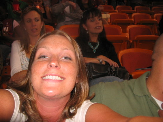 Woman with freckles smiles at camera in stadium seating, with other people behind her