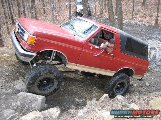 Red SUV with lifted suspension and large tires navigating rocky terrain. Another white SUV is visible in the background