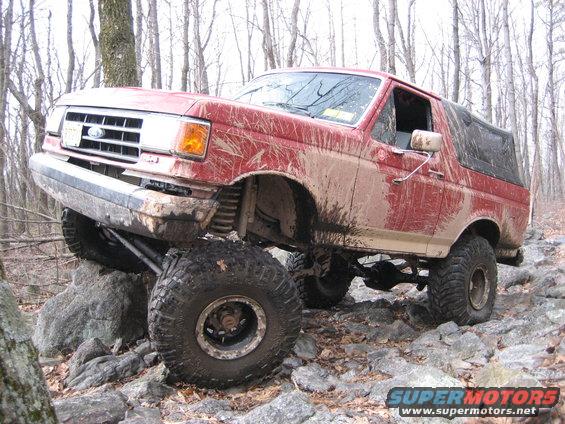 Mud-splattered red Ford Bronco on rocks, front-left tire elevated