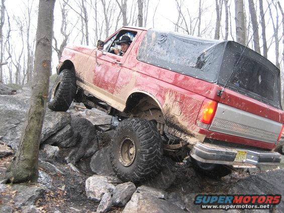 Red 1990 Ford Bronco climbing over large rocks on a trail. Driver wears a hat