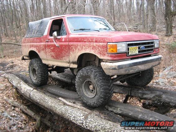 Red 1990 Ford Bronco with lifted suspension and off-road tires driving over logs