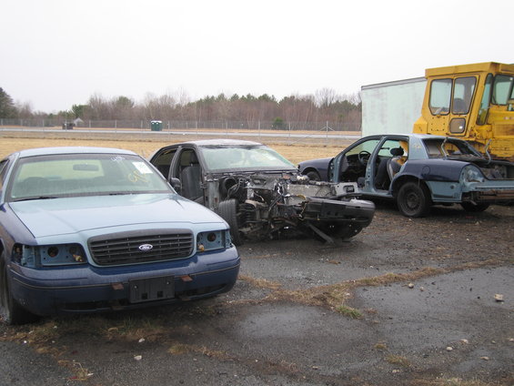 Three Ford Crown Victorias in a junkyard, two are heavily damaged. The front car is light blue, the middle car is dark blue and missing its front end. The rear car is dark blue and missing its rear end