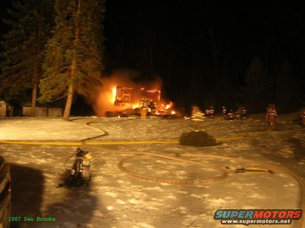 Burning structure at night with firefighters, hoses, and a fire extinguisher on snowy ground