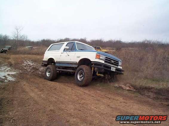 White and blue Ford Bronco with large tires on a dirt trail, 1987